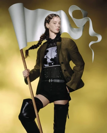 Model holding a large white flag banner wearing an olive blazer and black thigh-high boots against a warm yellow gradient background.