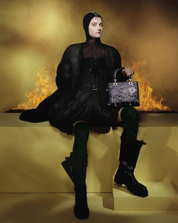 Model in black quilted coat and hood holding a handbag against a fiery backdrop for Dior Cruise 2025 campaign