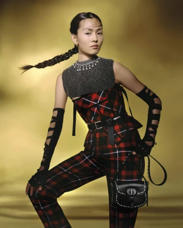 Model in a red and black plaid outfit holding a studded black handbag against a warm gradient background