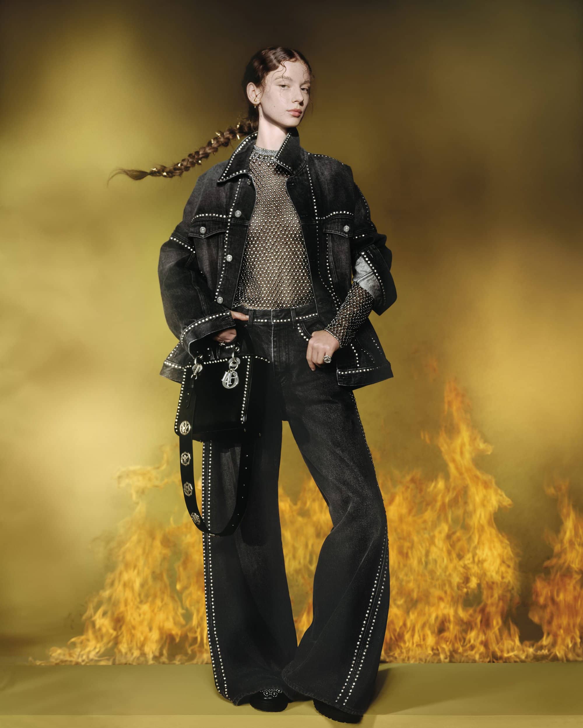 Model in embellished dark denim jacket and matching jeans holding a black handbag before a smoky yellow background with flames