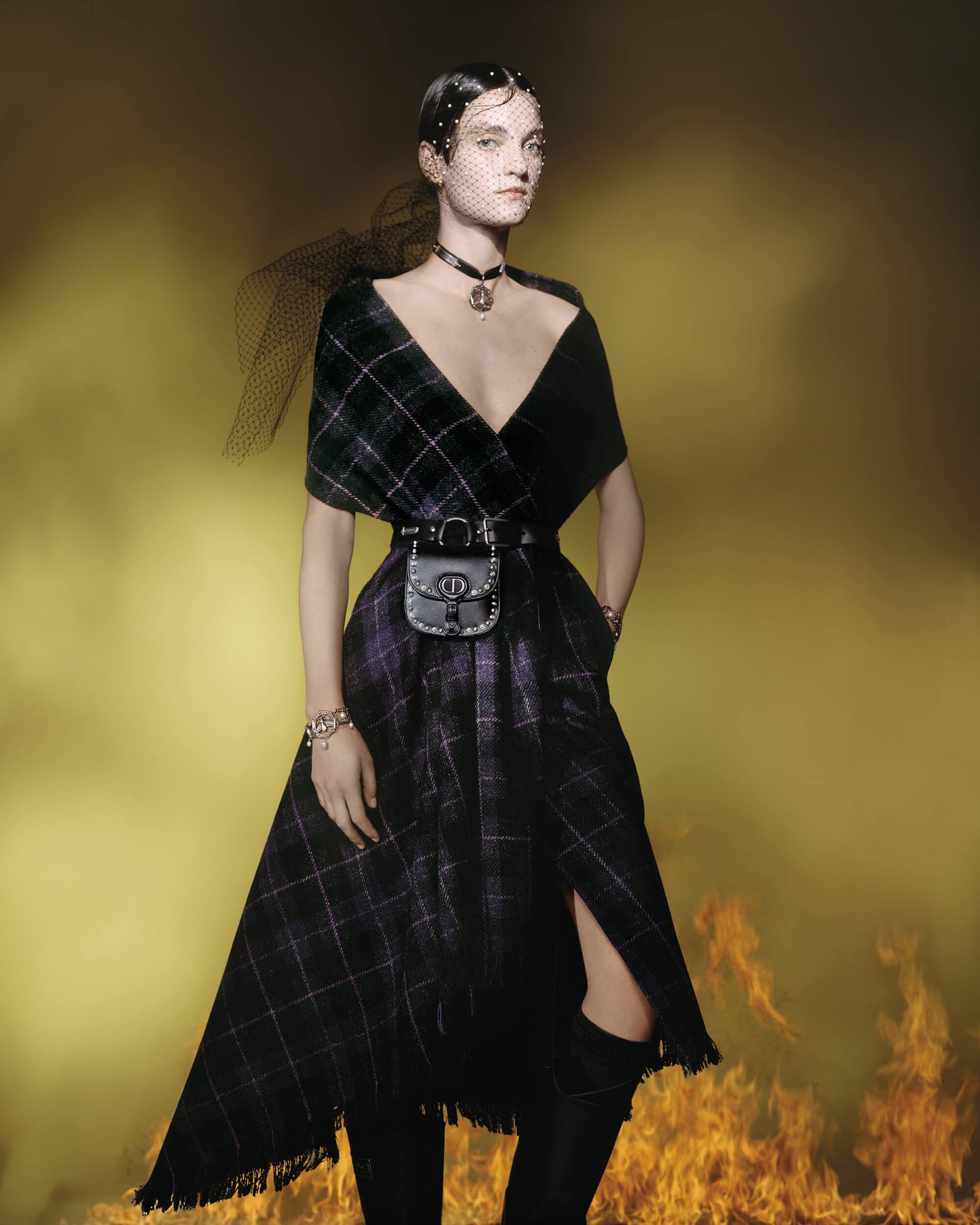 Model wearing a dark plaid dress and veil against a smoky, yellow-lit background for the Dior Cruise 2025 campaign.