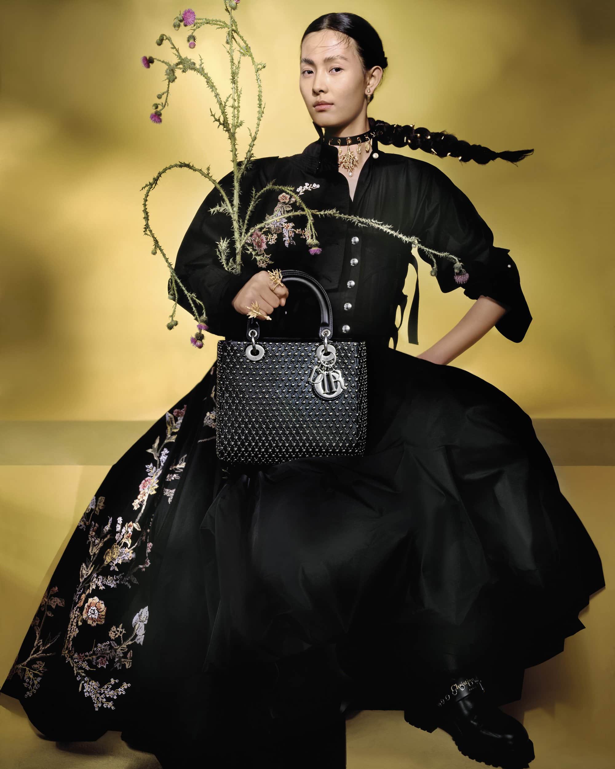 Model in black dress holding a structured handbag and flowering branches against a warm yellow gradient background.