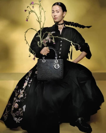 Model in black dress holding a structured handbag and flowering branches against a warm yellow gradient background.