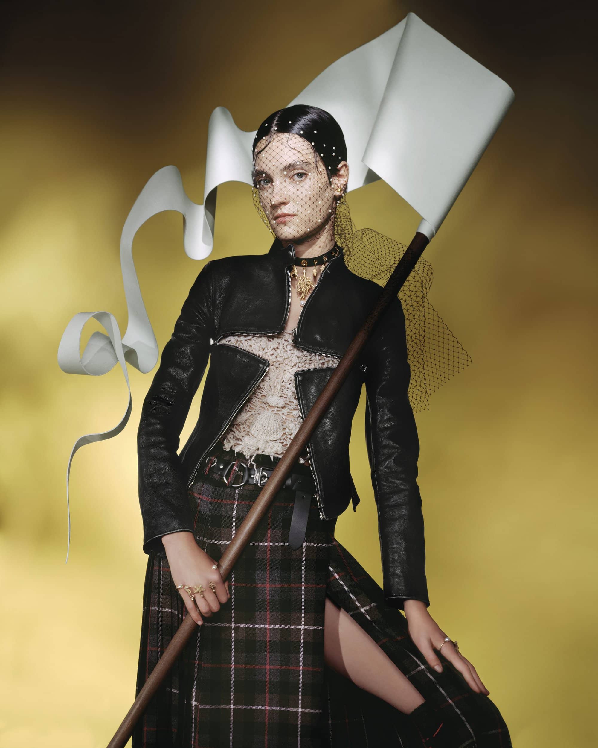 Model holding a staff with a fluttering white banner wearing a black leather jacket and plaid skirt against a yellow background.