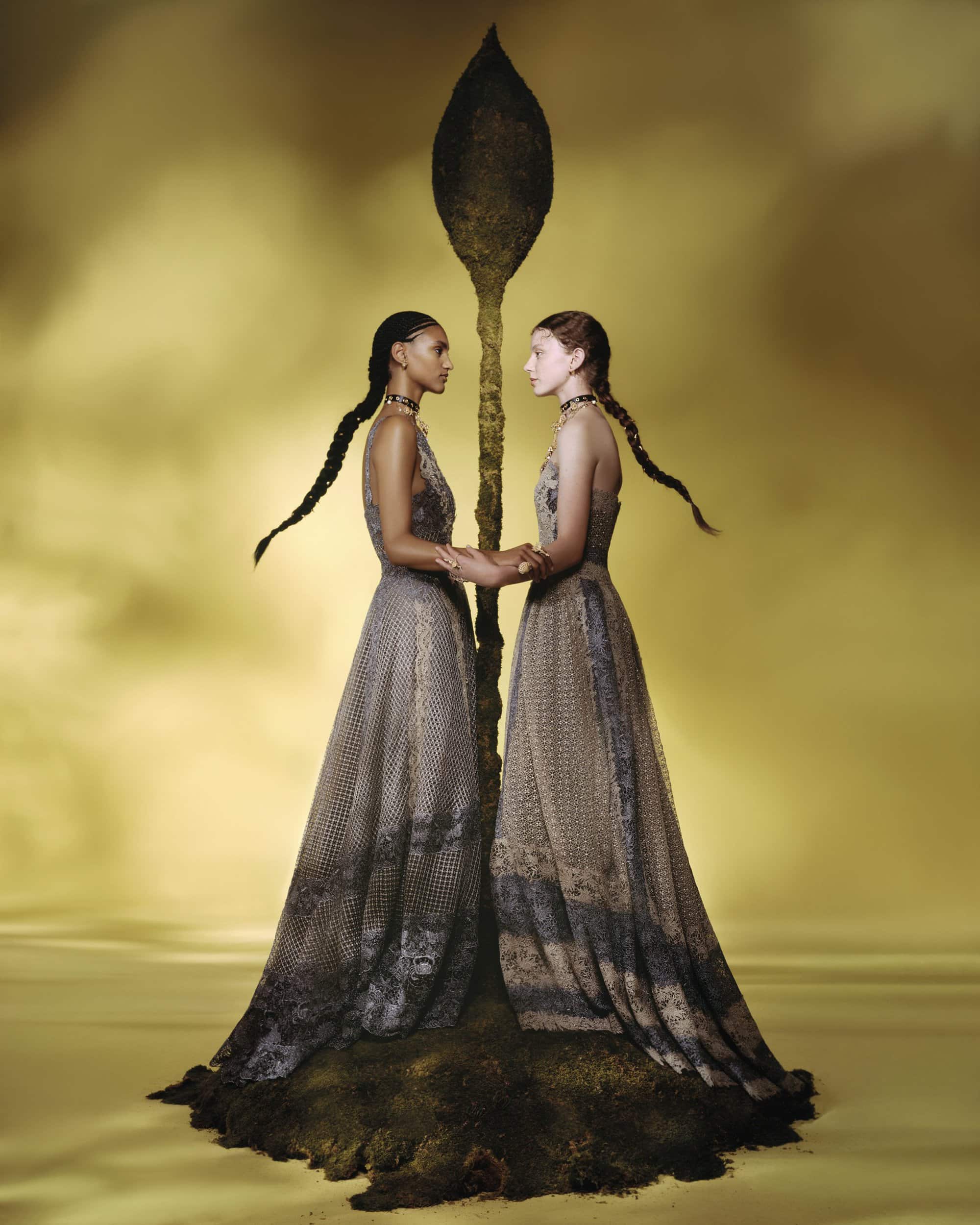 Two female models in long patterned dresses holding hands before an organic sculpture against a warm hazy backdrop.