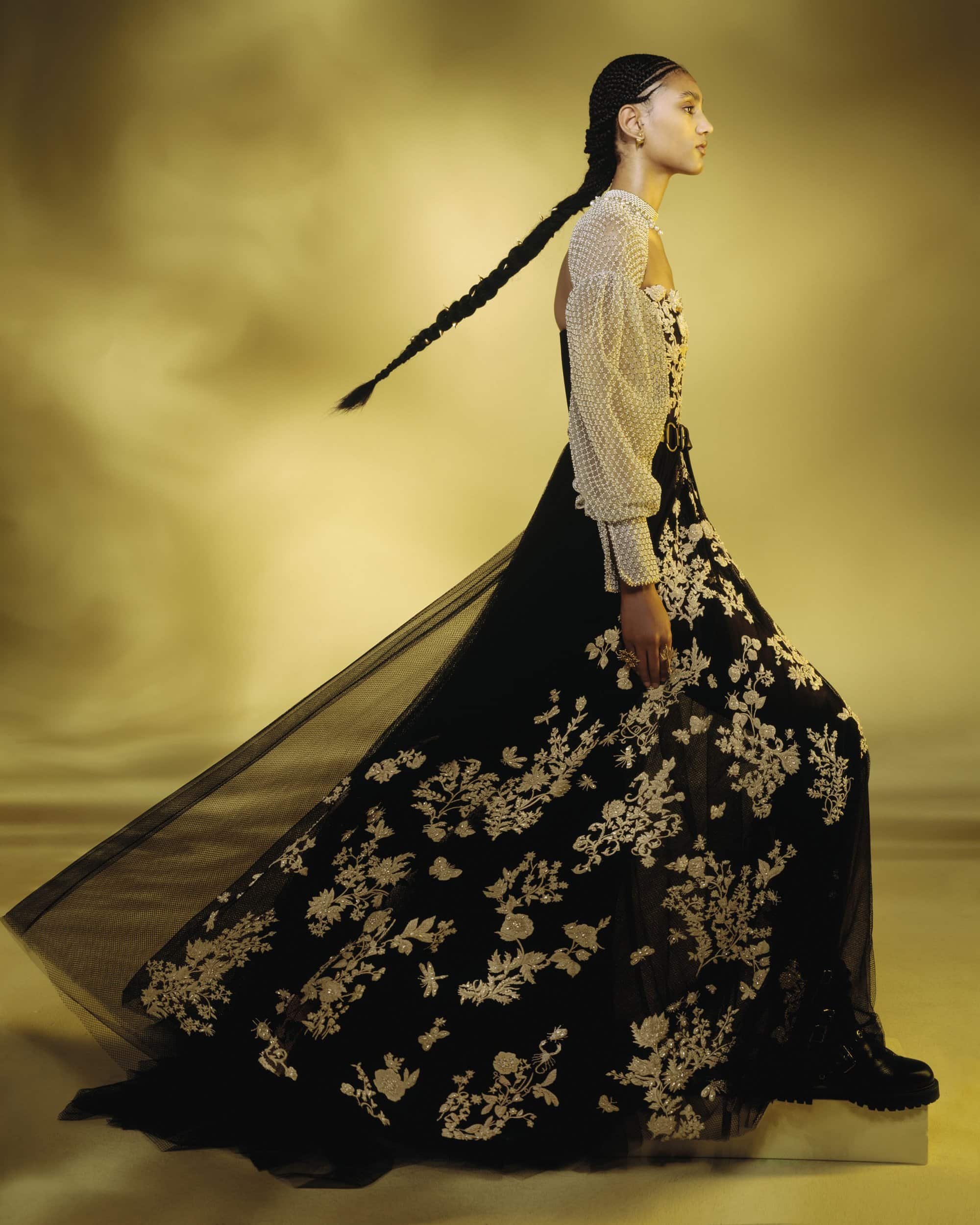 Model in a black and white embroidered gown with a long braid against a soft warm gradient background.