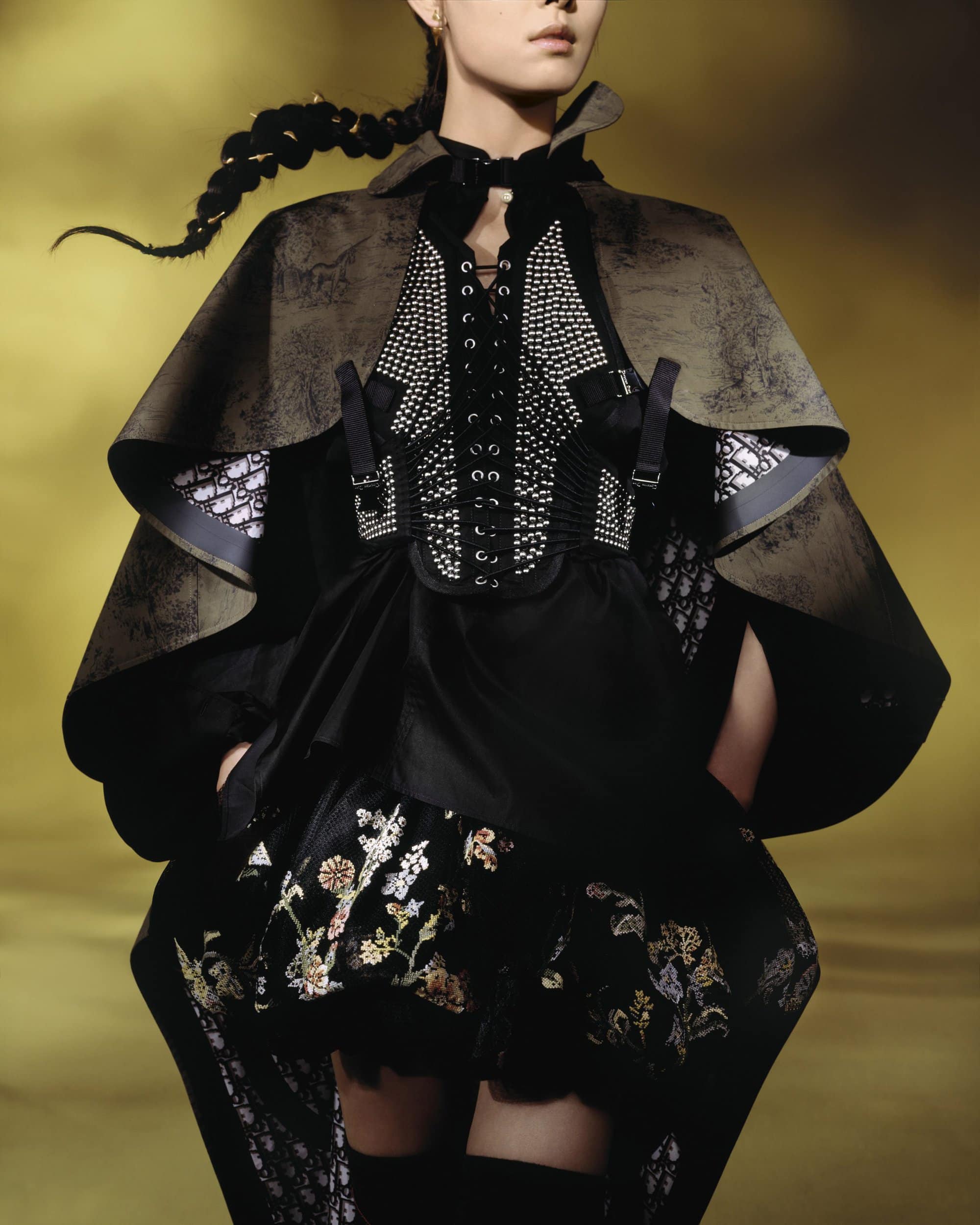 Model wearing a dark cape, laced corset with studs, and embroidered skirt against a warm background.