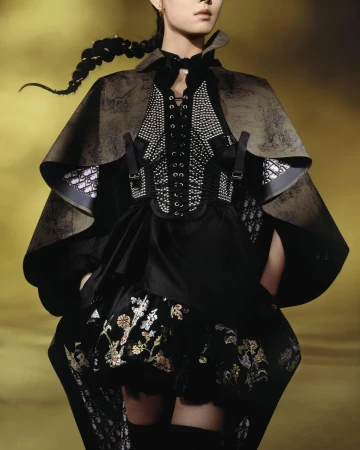 Model wearing a dark cape, laced corset with studs, and embroidered skirt against a warm background.