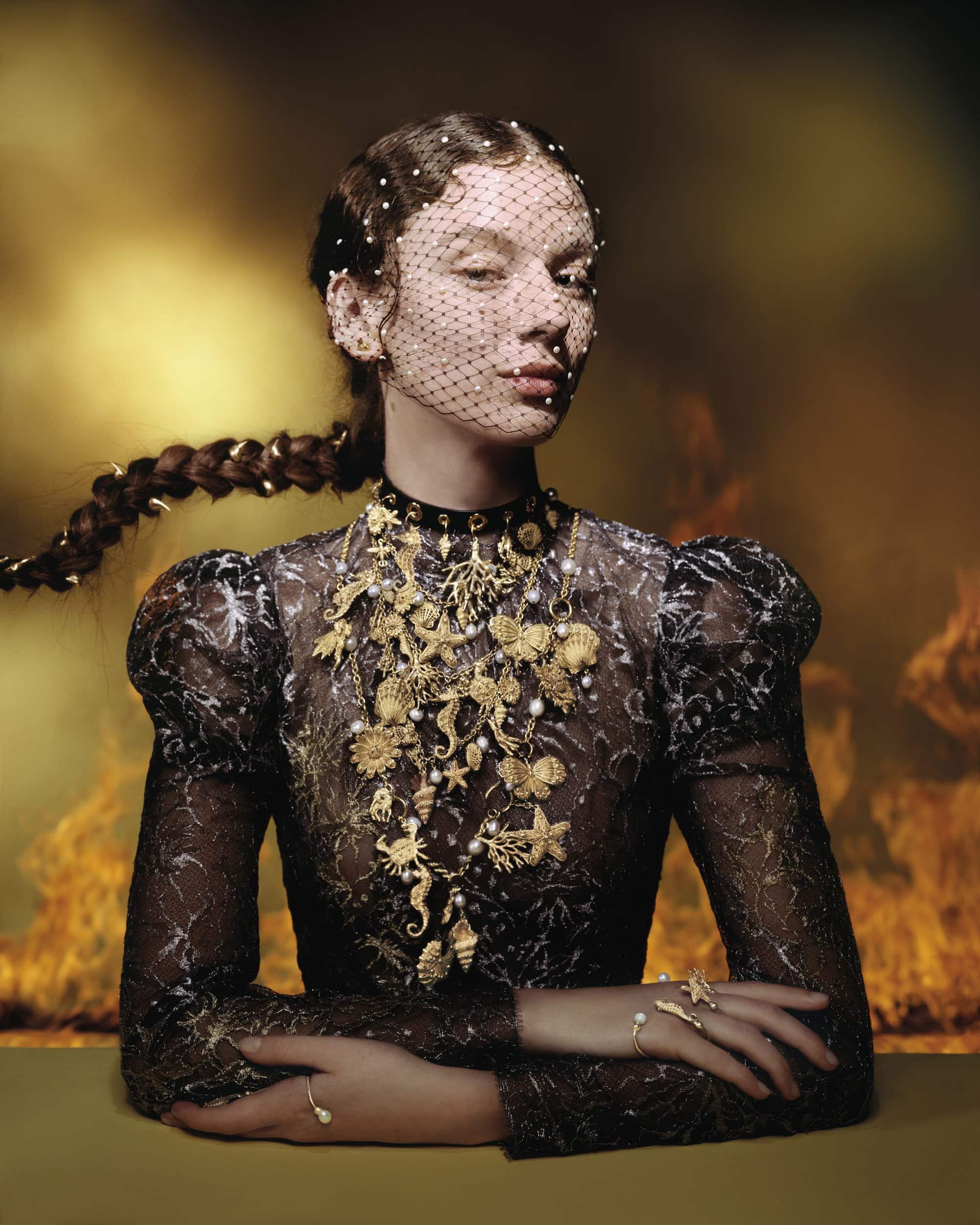 Model wearing a black lace top with ornate gold jewelry and a beaded face veil against a moody backdrop