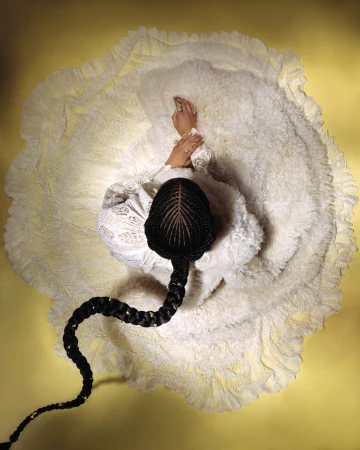 Overhead shot of a model in a white ruffled dress with a long braided hairstyle against a yellow backdrop.