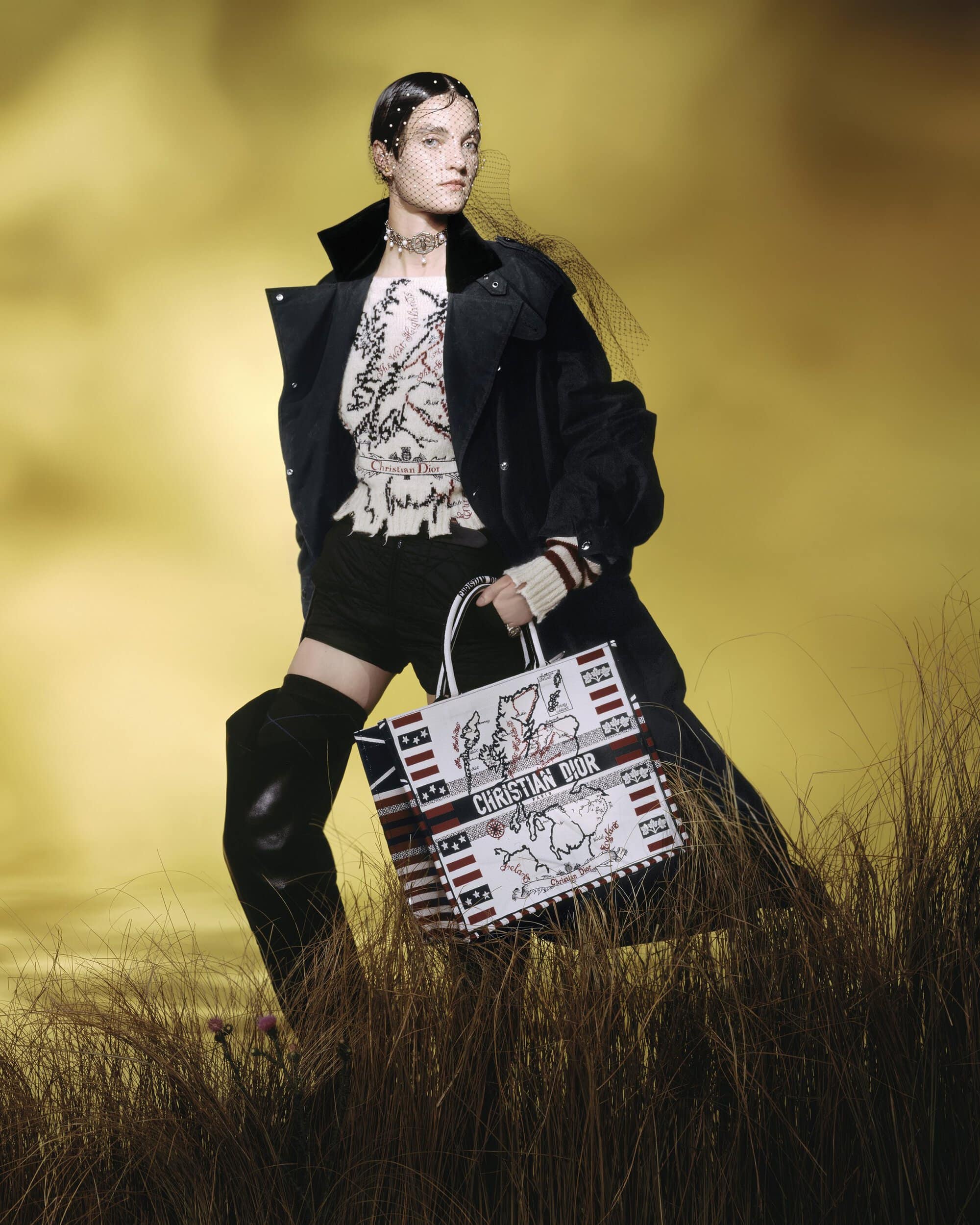 Model wearing dark coat and knee-high boots holding patterned tote bag standing in tall grass against a yellow sky.