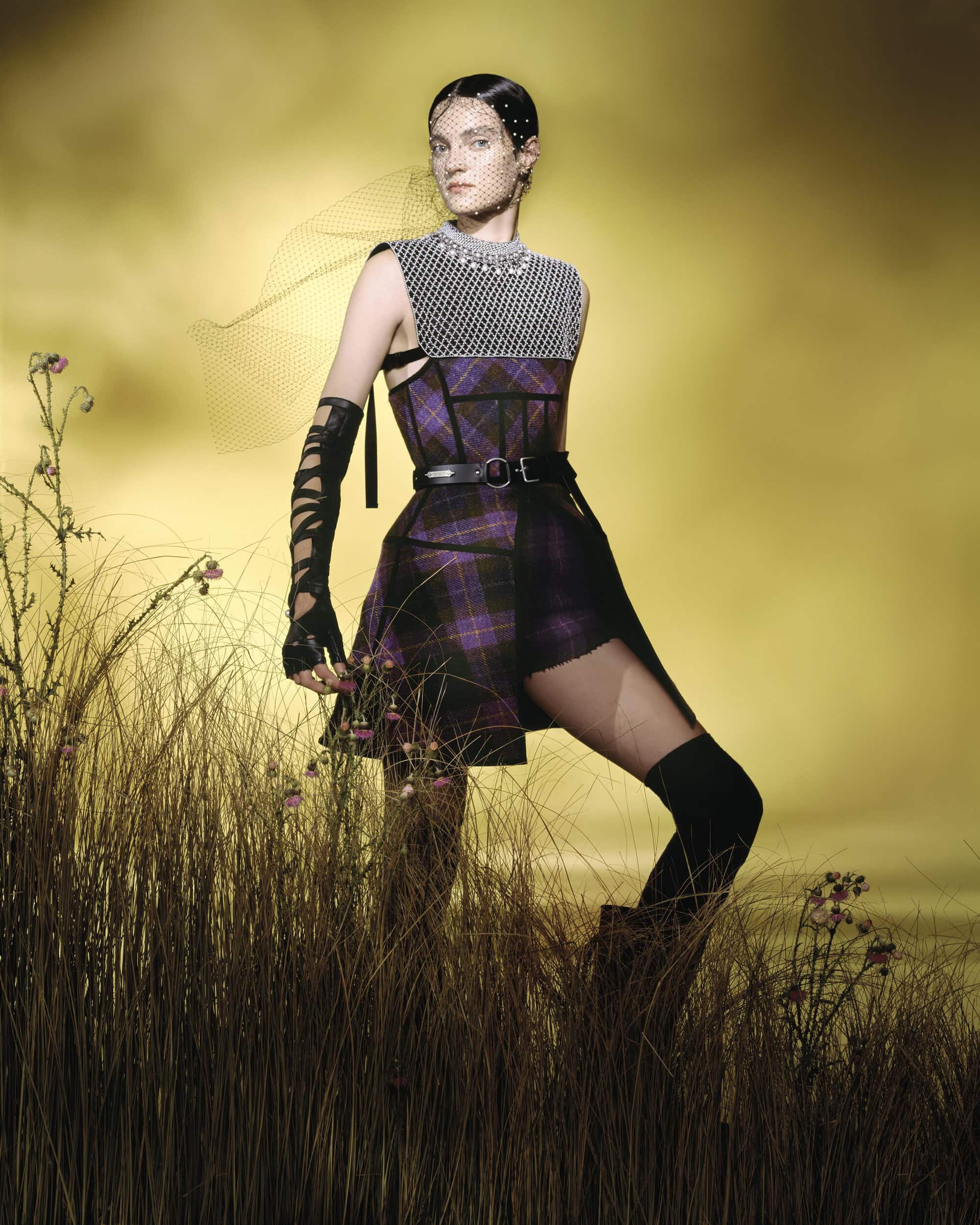 Model wearing a plaid dress and chainmail top standing in tall grass against a dramatic yellow sky