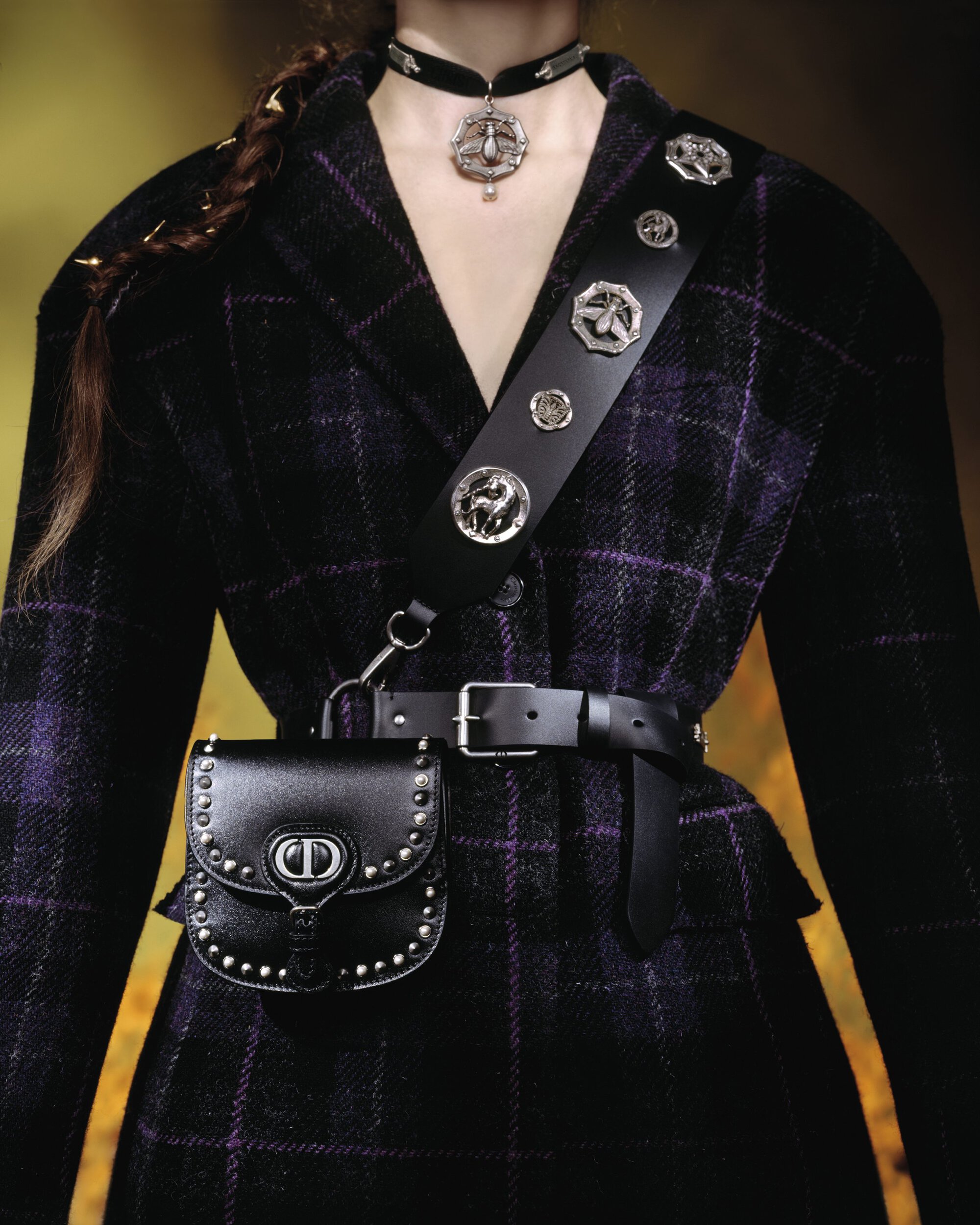 Model wearing a dark plaid jacket, black leather bag, studded strap, and choker necklace for Dior Cruise 2025 campaign.