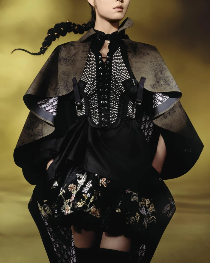 Model wearing a dark cape, laced corset with studs, and embroidered skirt against a warm background.