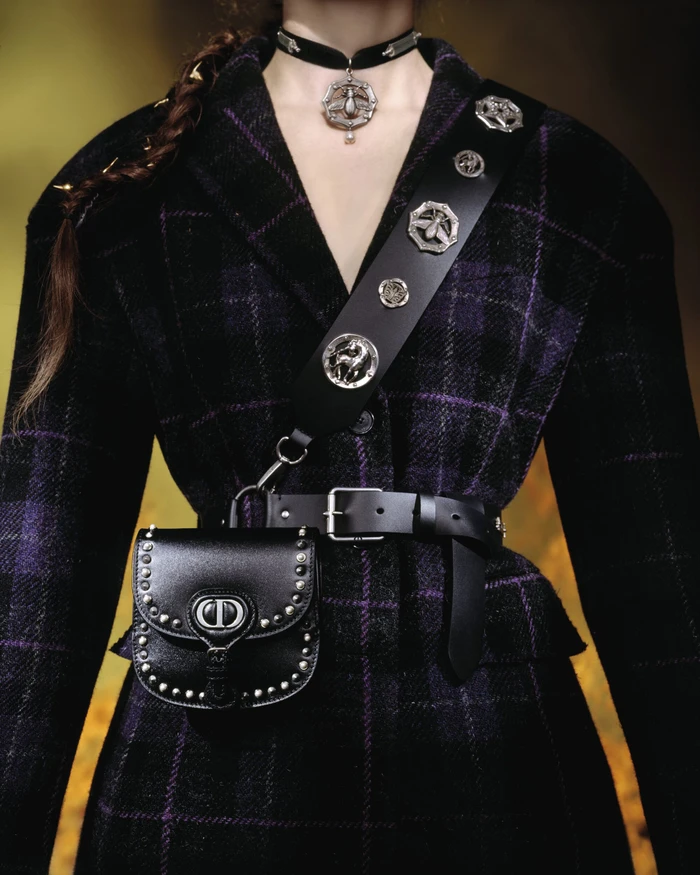 Model wearing a dark plaid jacket, black leather bag, studded strap, and choker necklace for Dior Cruise 2025 campaign.