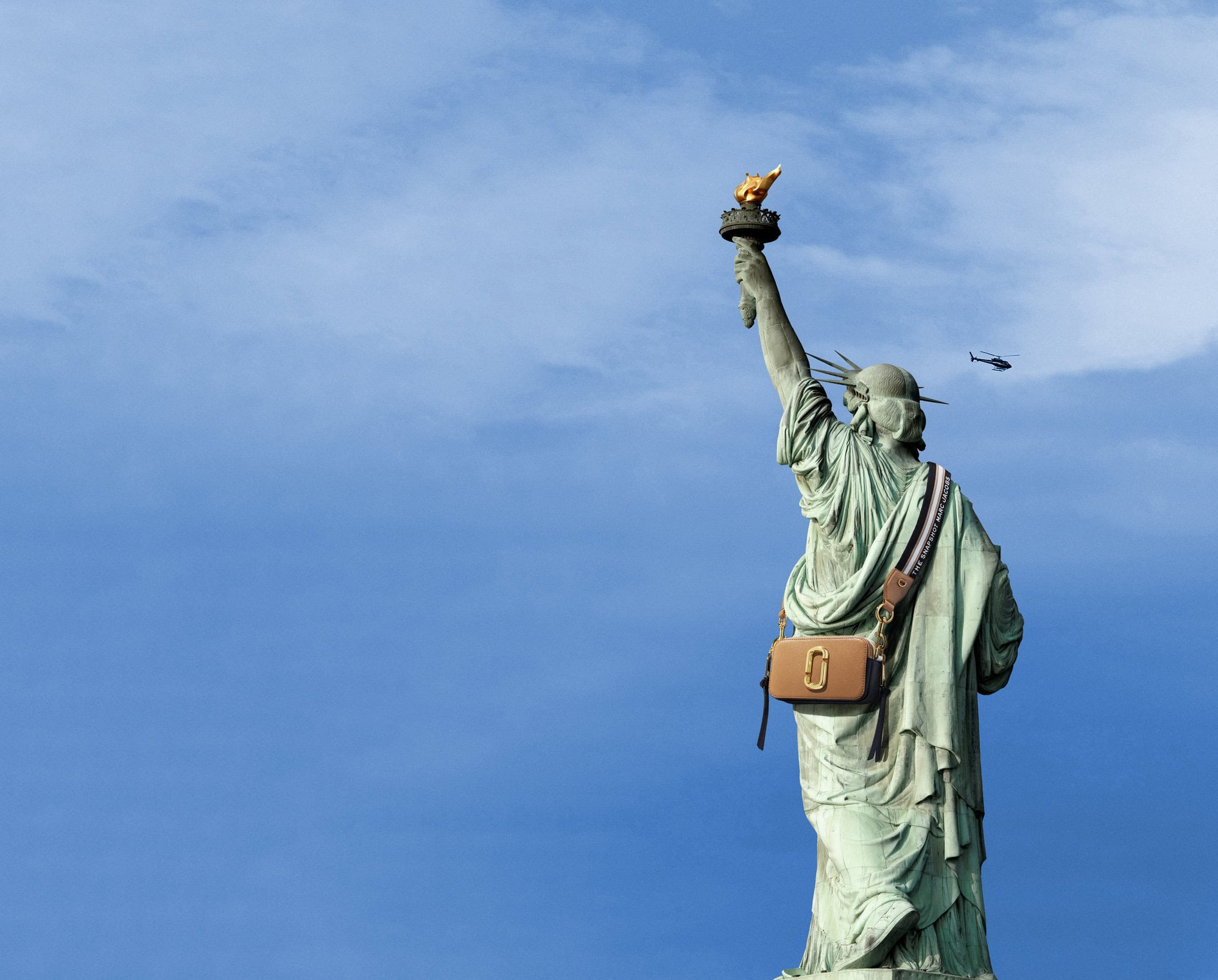 Statue of Liberty from behind wearing a tan crossbody handbag against a clear blue sky with a small helicopter visible in the distance