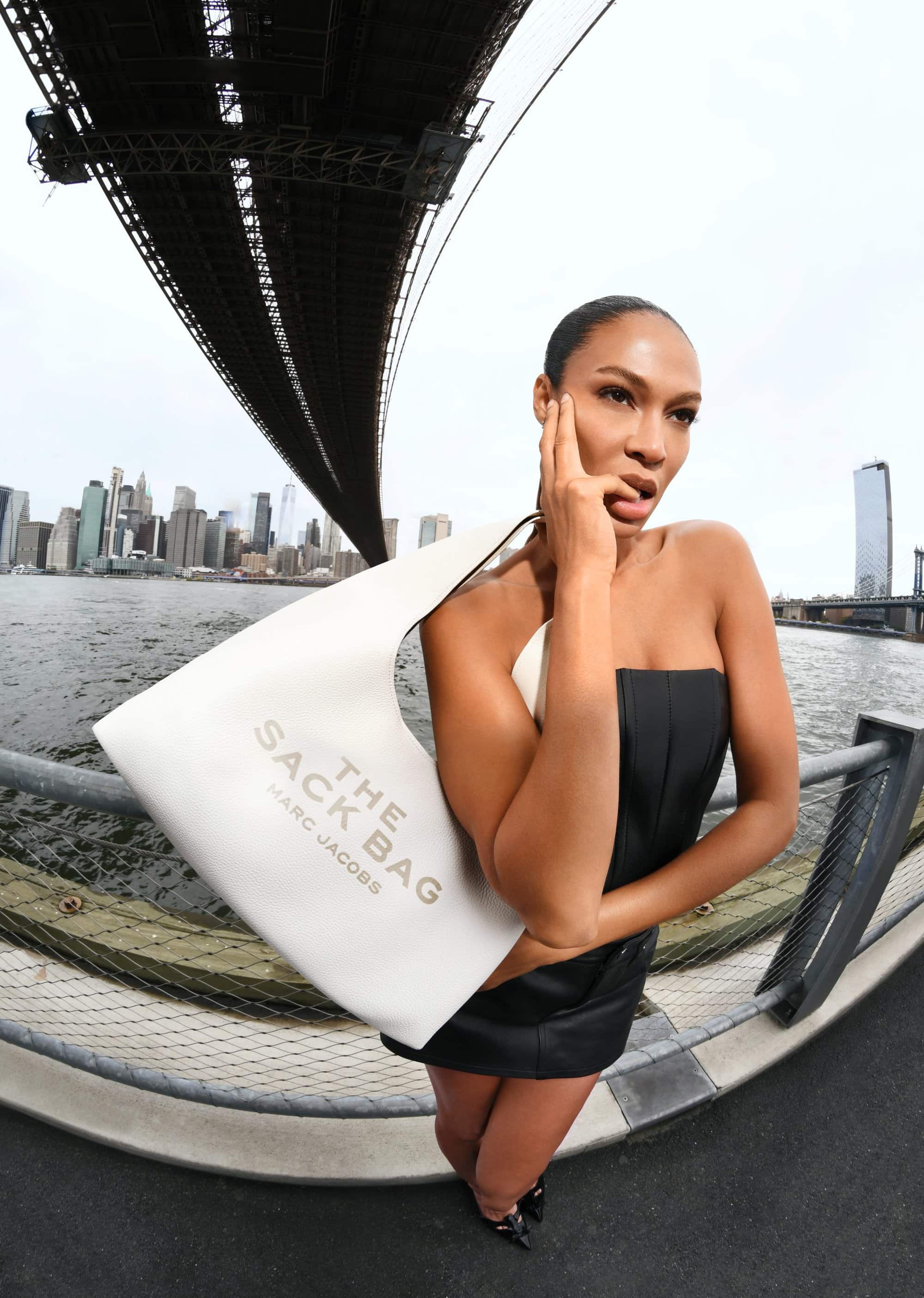 Model Joan Smalls holding a white shoulder bag outdoors against a bridge and city skyline.