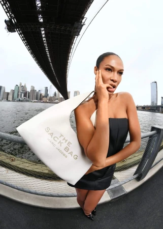 Model Joan Smalls holding a white shoulder bag outdoors against a bridge and city skyline.