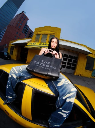 Model seated on the roof of a yellow cab holding a black tote bag in front of a garage building.