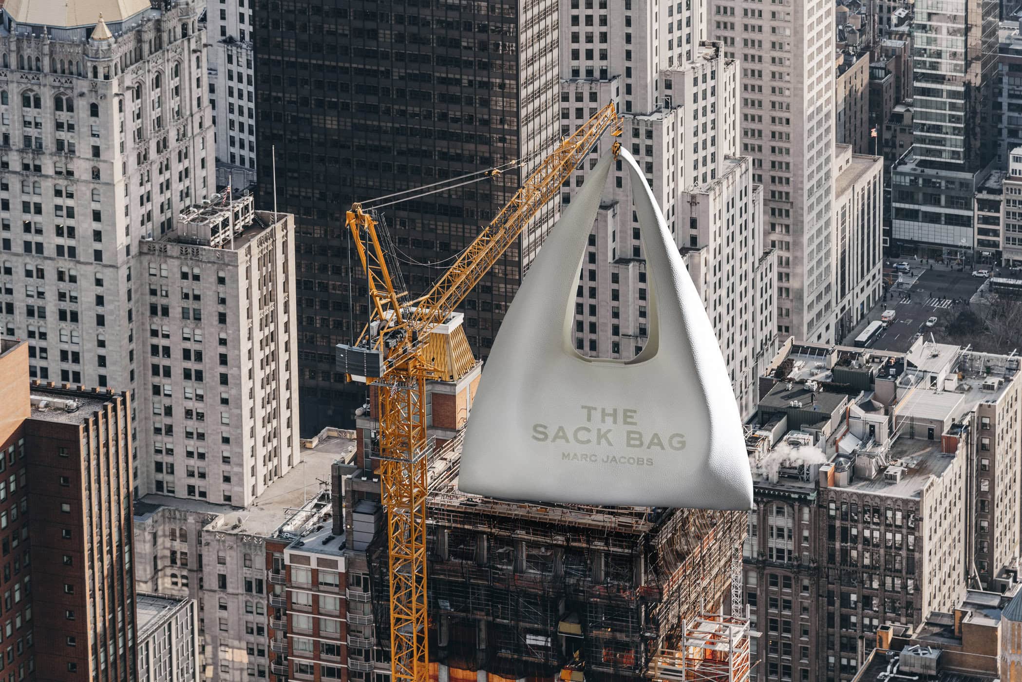 A massive white Marc Jacobs handbag hoisted in the air by a yellow construction crane above New York City skyscrapers.