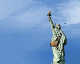 Statue of Liberty from behind wearing a tan crossbody handbag against a clear blue sky with a small helicopter visible in the distance
