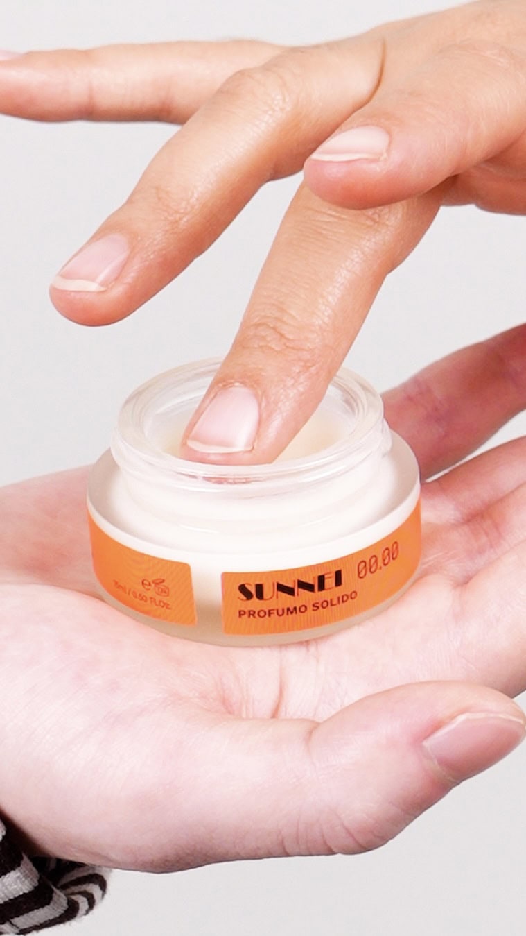 Close-up of a person holding a small white cosmetic jar with an orange label on an open palm and touching the product