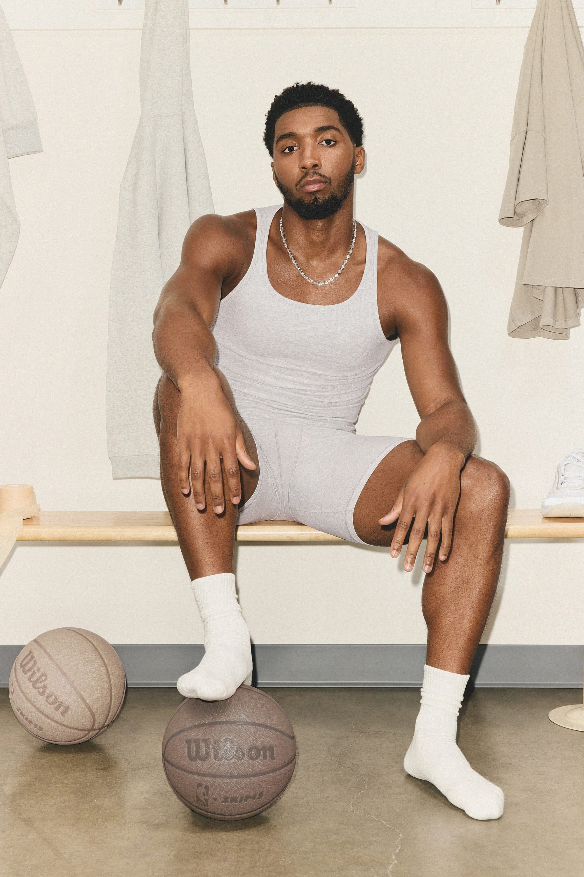 Donovan Mitchell seated on a wooden bench in a locker room wearing a grey tank top and shorts, with basketballs on the floor.