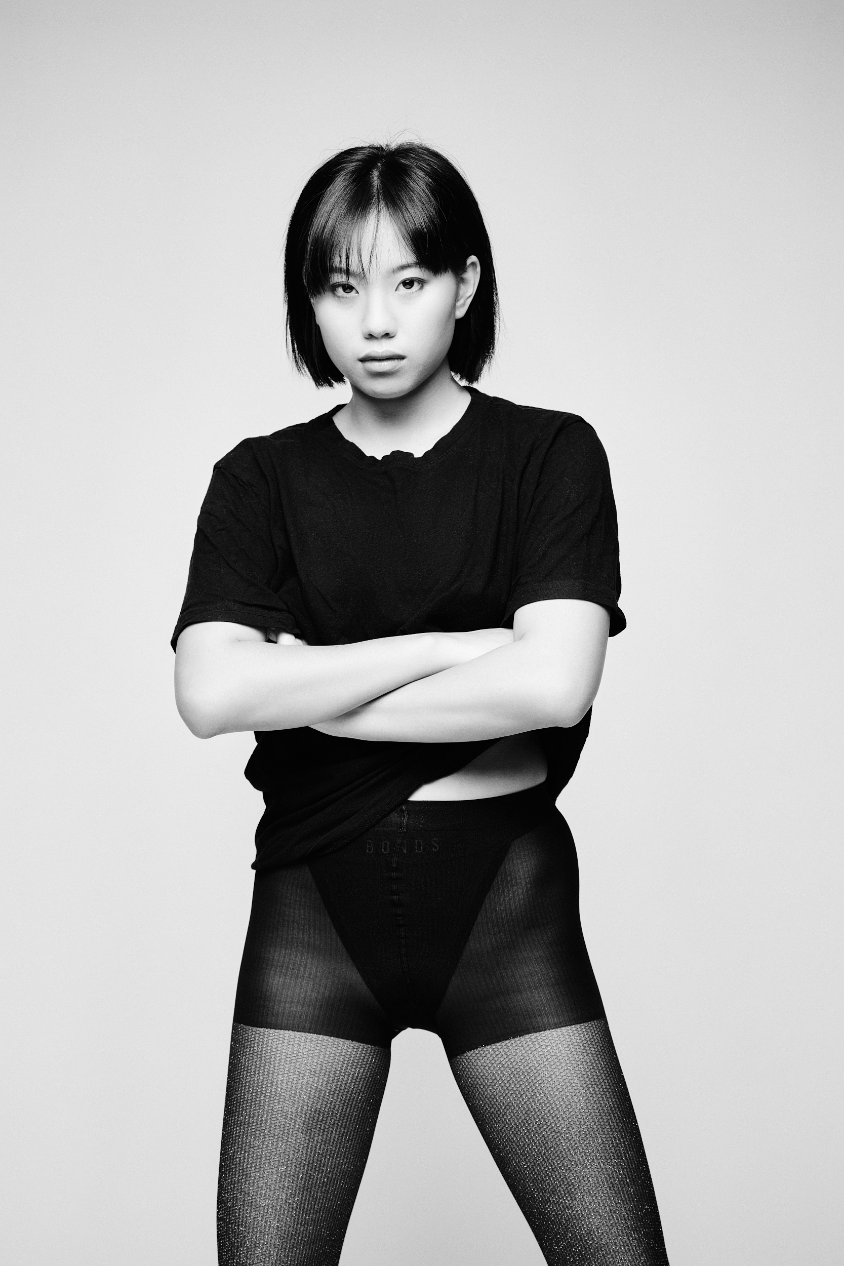 Black and white portrait of a confident young woman with short dark hair, wearing a dark t-shirt and textured tights, arms crossed.