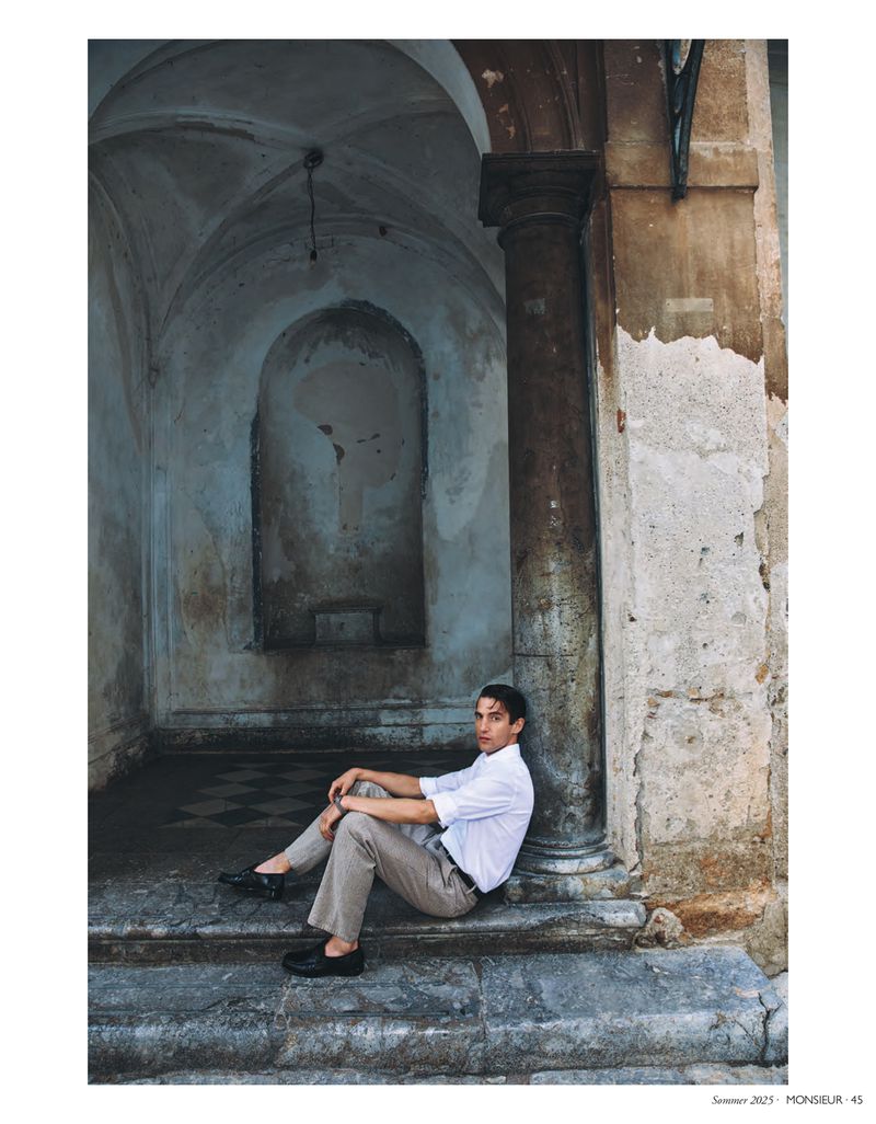 Young man sitting on stone steps with a pensive expression near a column. He wears a white shirt and khaki pants in an old building.