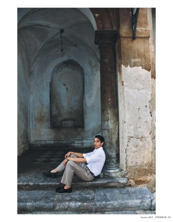 Young man sitting on stone steps with a pensive expression near a column. He wears a white shirt and khaki pants in an old building.