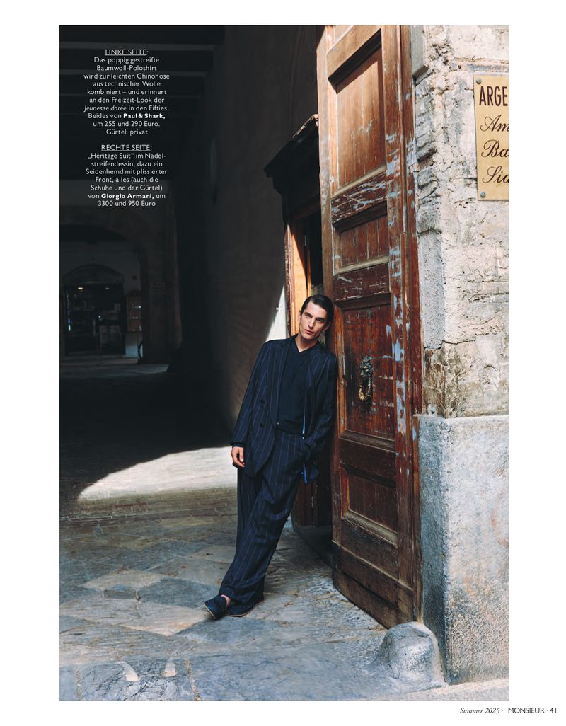 A fashionable adult man wearing a pinstripe suit and suspenders poses outdoors in an alleyway leaning against an old wooden door.