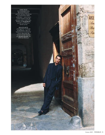 A fashionable adult man wearing a pinstripe suit and suspenders poses outdoors in an alleyway leaning against an old wooden door.