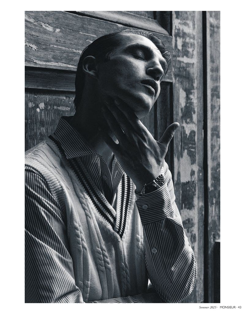 Black and white portrait of an adult male with closed eyes, striped shirt, and cable knit sweater in front of a weathered door.