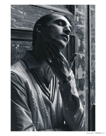 Black and white portrait of an adult male with closed eyes, striped shirt, and cable knit sweater in front of a weathered door.