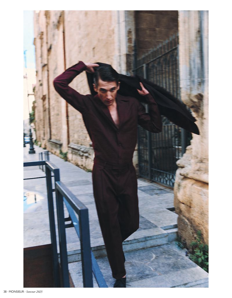 An adult male fashion model in a maroon jumpsuit, walking toward the camera, removing his jacket outdoors against a sandstone building.