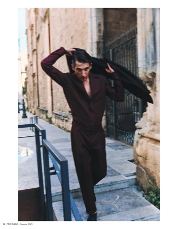 An adult male fashion model in a maroon jumpsuit, walking toward the camera, removing his jacket outdoors against a sandstone building.