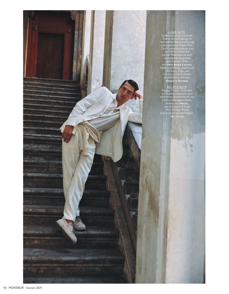 A relaxed, full-length portrait of a young adult male in a white linen suit, posing on stone steps near a weathered building exterior.