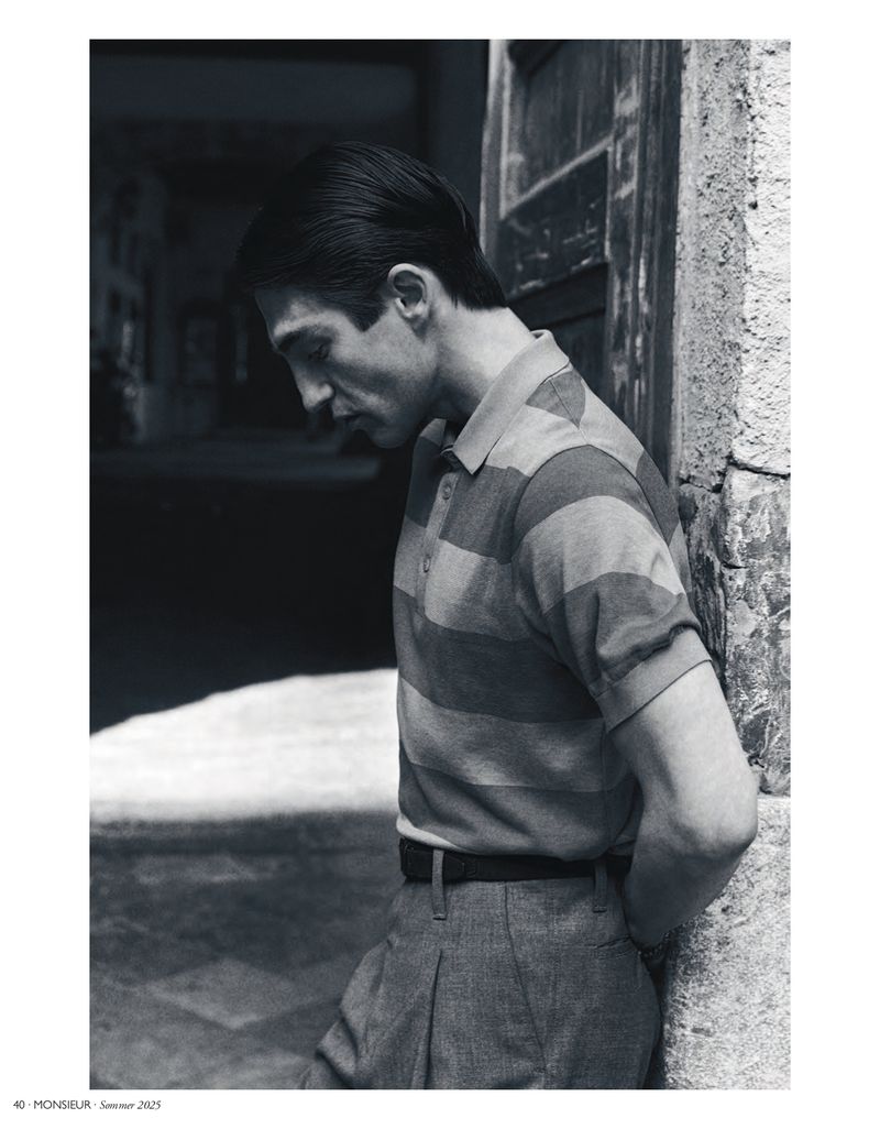 Monochrome portrait of a stylish man in a striped collared shirt leaning against a wall outside, with a blurred background.