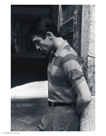 Monochrome portrait of a stylish man in a striped collared shirt leaning against a wall outside, with a blurred background.