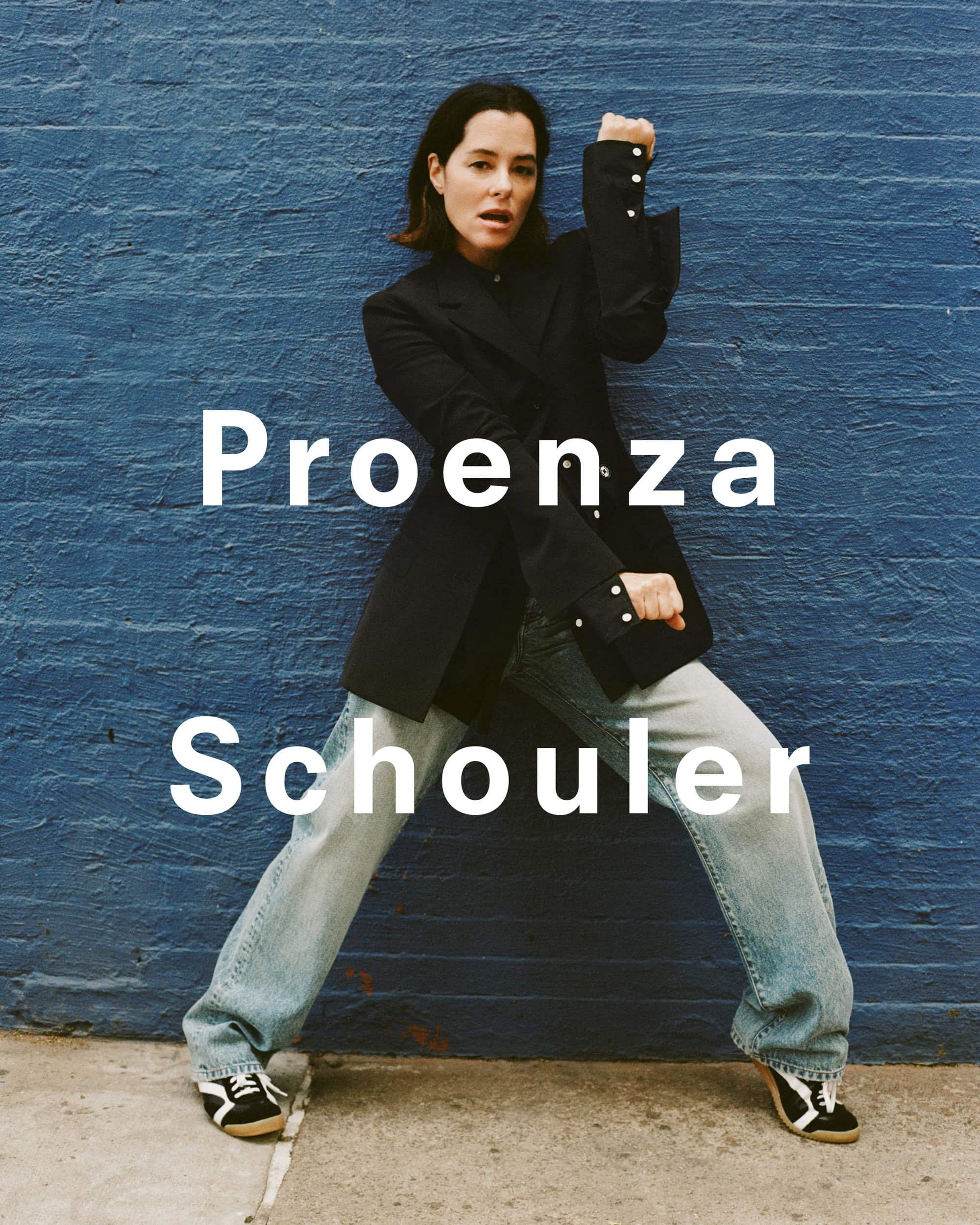 Parker Posey posing in a black blazer and blue jeans against a textured blue brick wall for Proenza Schouler
