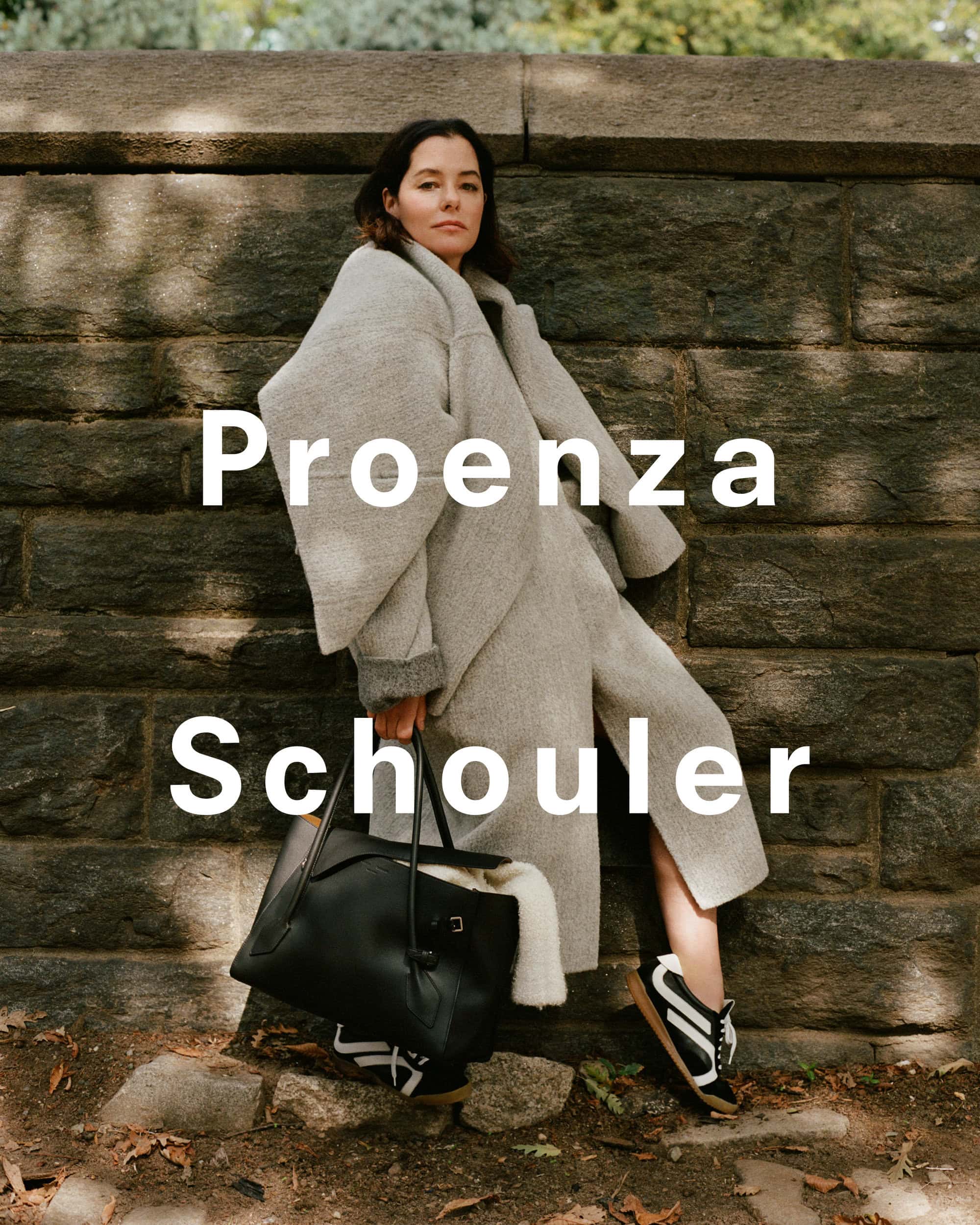 Parker Posey leaning against a stone wall wearing a grey coat and holding a large black leather handbag