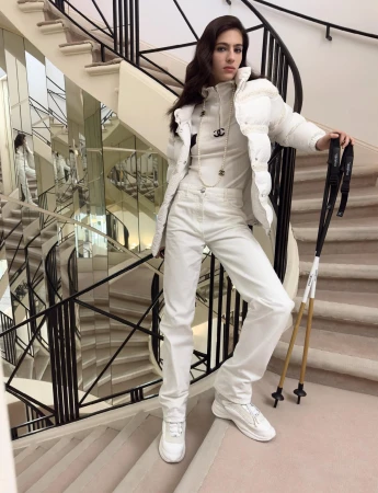 Model Loli Bahia standing on a curved staircase holding ski poles, wearing a white puffer jacket and white trousers.