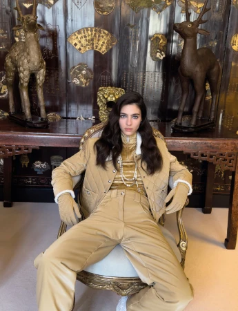 Model Loli Bahia seated in an ornate chair wearing a quilted beige jacket and trousers for Chanel Coco Neige 2024 ad campaign.