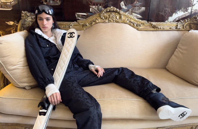 Model Loli Bahia seated on an ornate sofa holding white Chanel skis, wearing a black ski suit and ski goggles