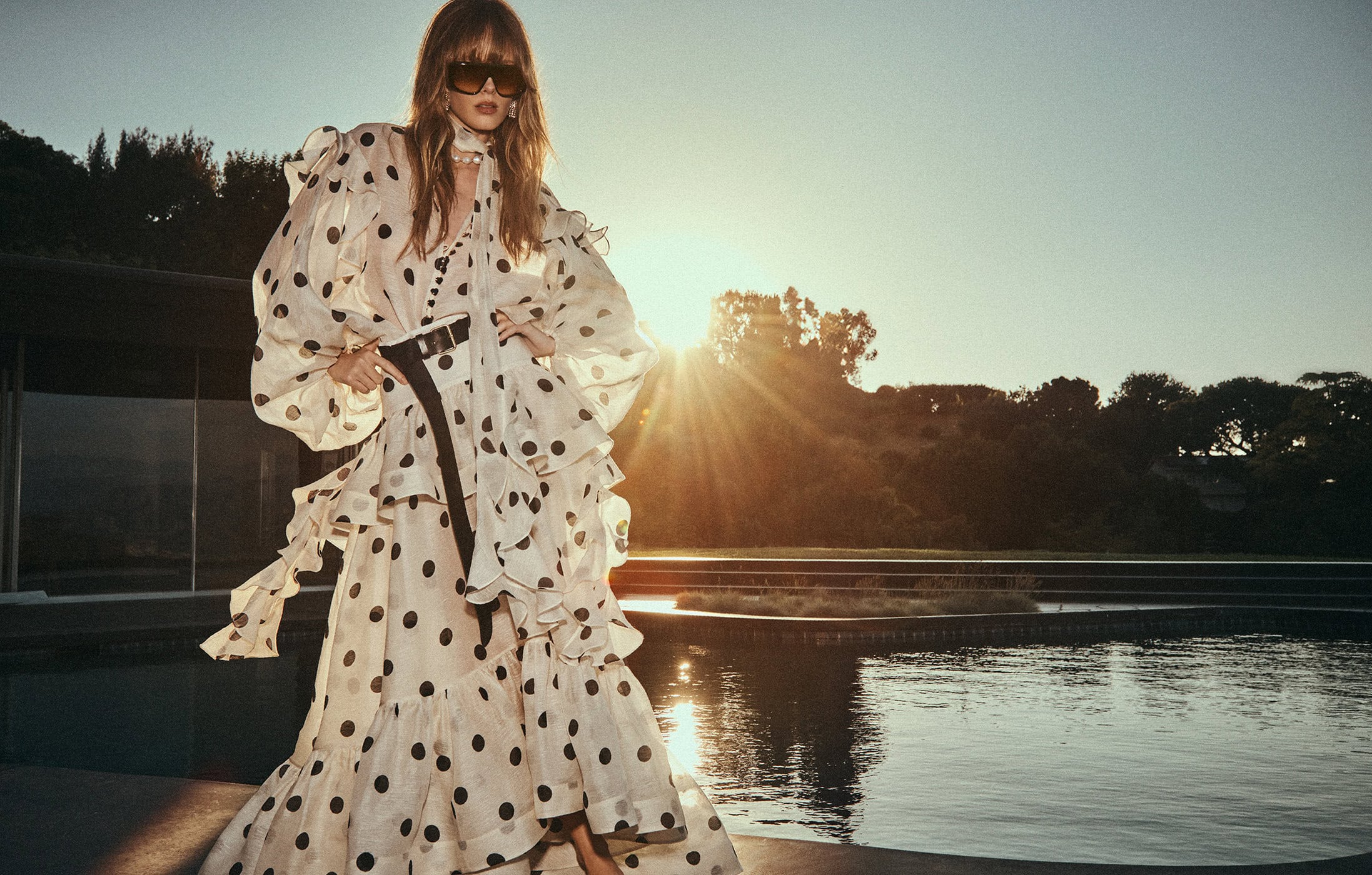 Model in a long polka dot dress standing by a swimming pool at sunset for Zimmermann Resort 2025 campaign.
