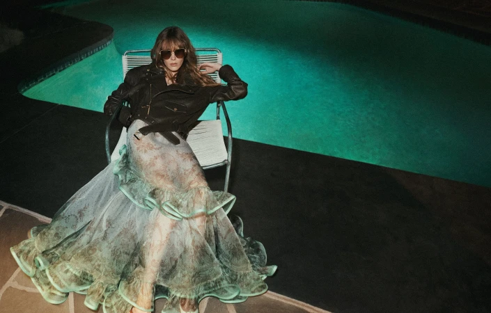 Model wearing dark sunglasses, a black leather jacket, and a ruffled patterned gown seated on a chair by a glowing green pool at night.