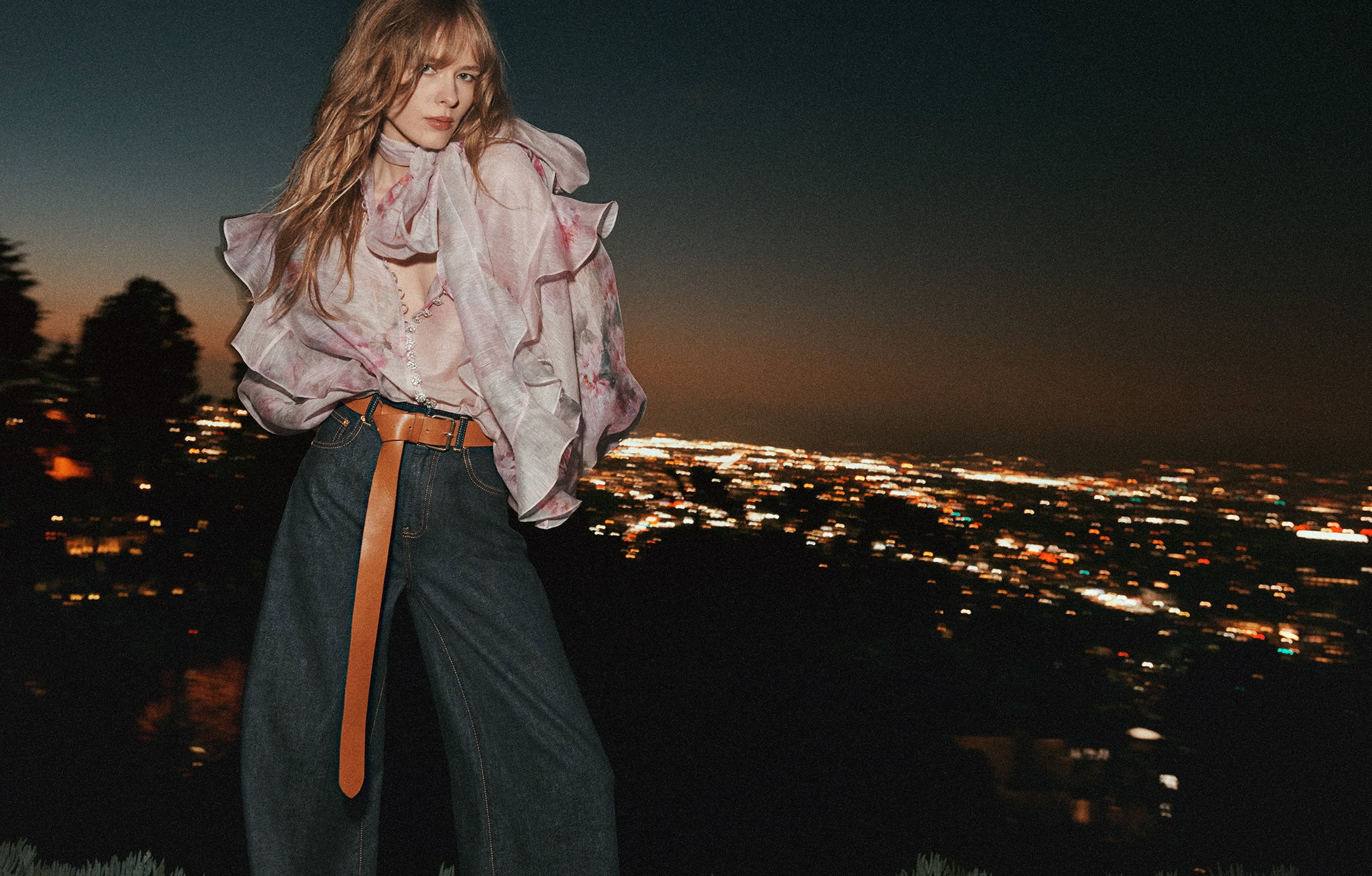 Model in ruffled floral blouse and denim jeans posing against a glowing night city skyline backdrop