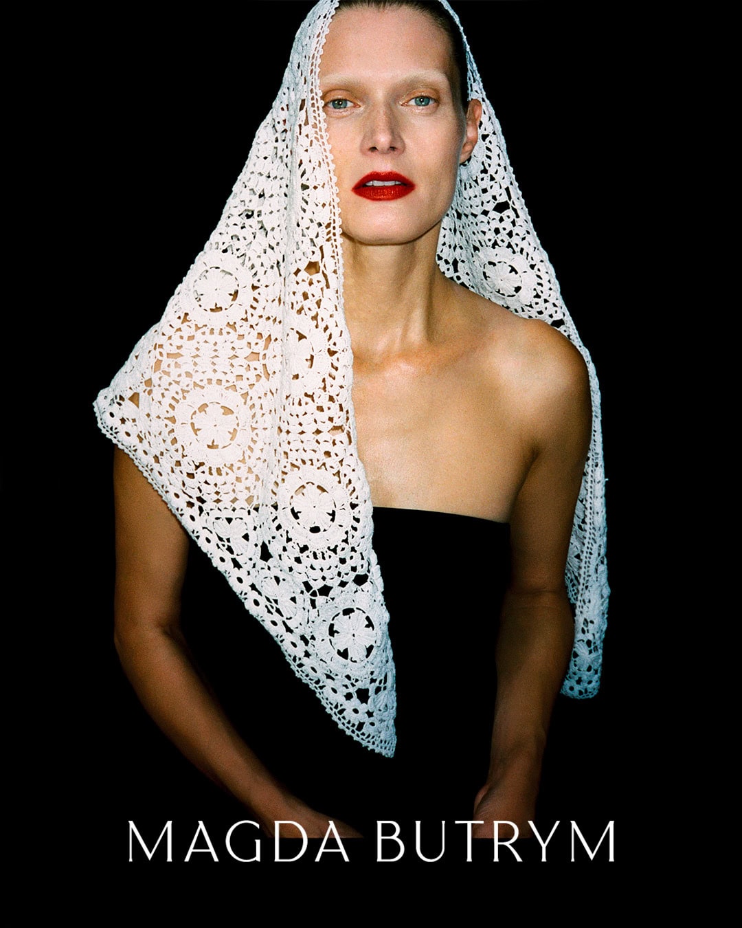 Model wearing a white lace head covering and bold red lipstick against a solid black background for Magda Butrym campaign