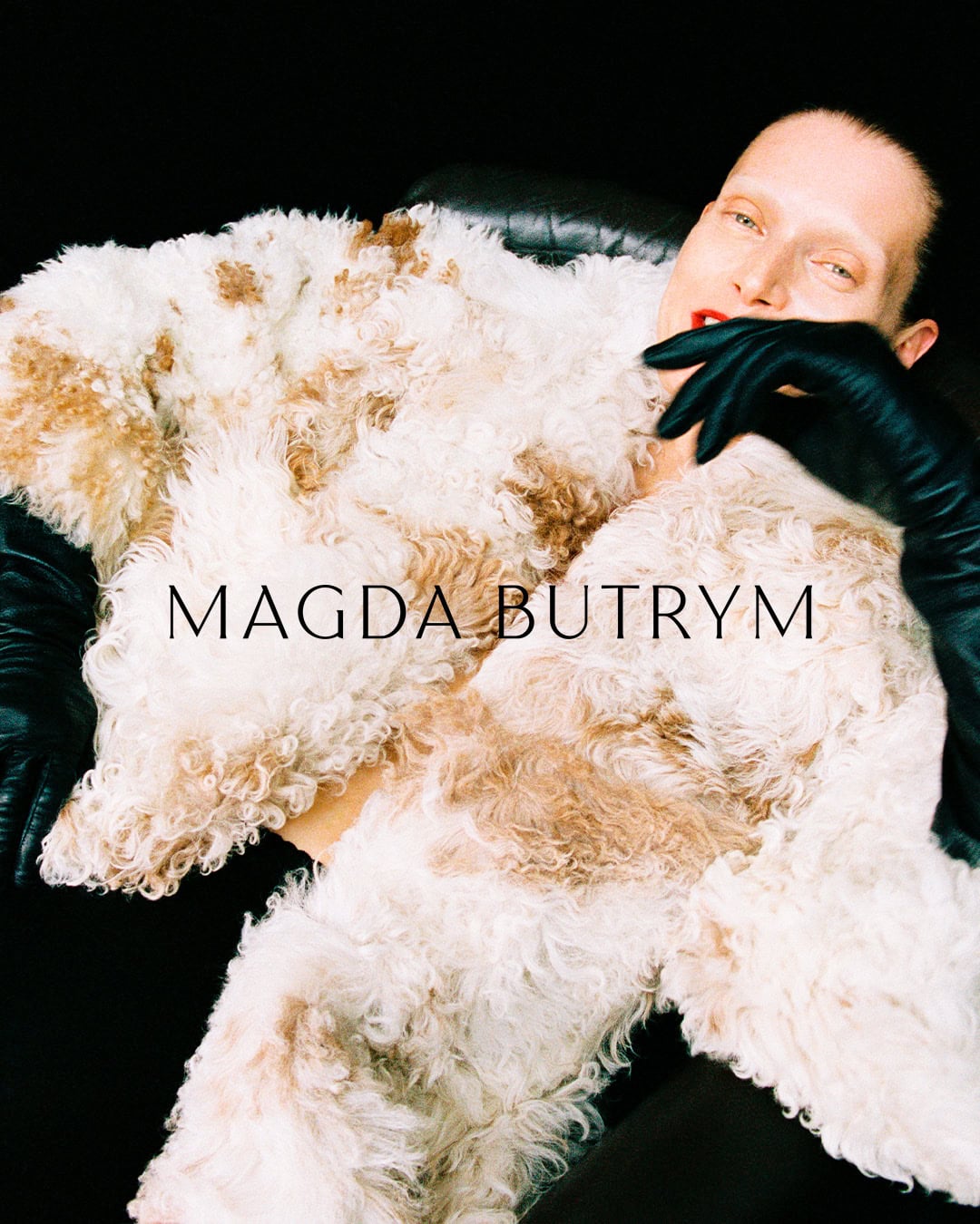 Model wearing a textured patterned coat and long black leather gloves against a dark background with brand text