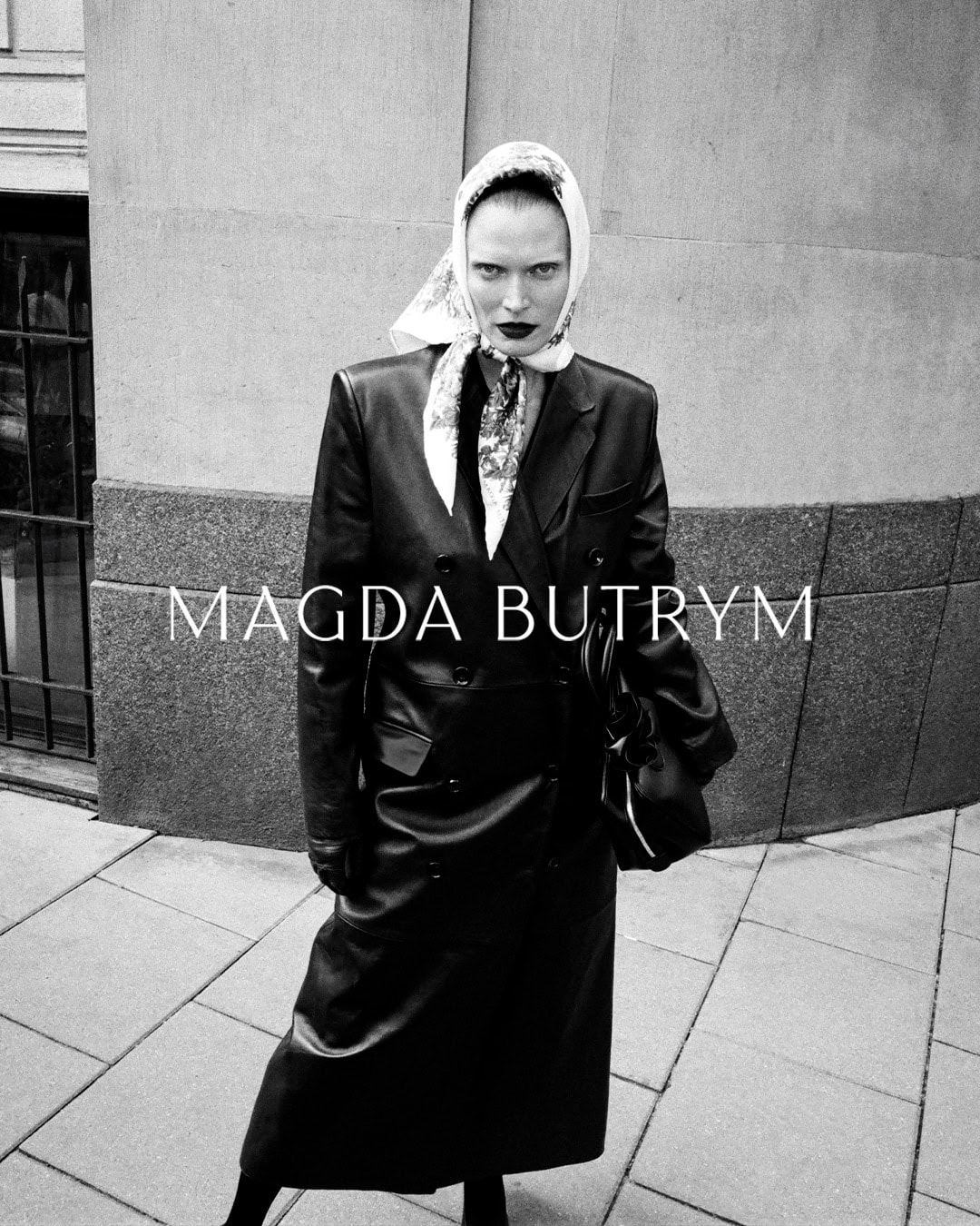 Model wearing a leather coat and headscarf holding a handbag on a city street for Magda Butrym